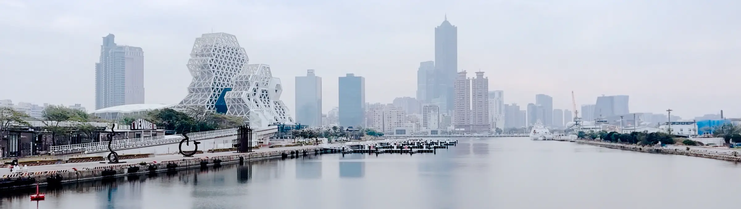 Panoramic black and white photo of the Kaohsiung cityscape, with the Kaohsiung Music Center in the foreground, and skyscrapers including 85 Sky Tower across the harbor in the distance.